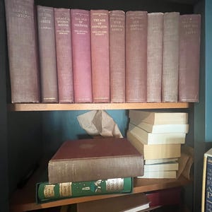 May include: A bookshelf filled with vintage books. The spines are various shades of red and brown, with titles visible. Some books are stacked on the shelves. The overall aesthetic suggests a classic or antique theme.