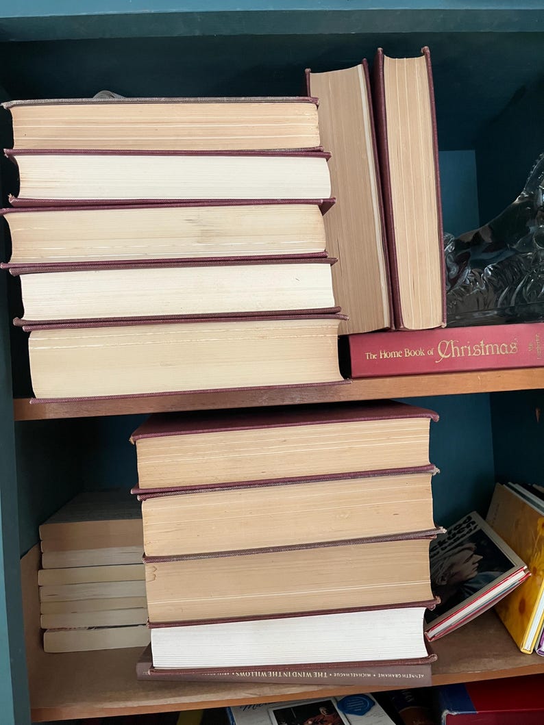 May include: A collection of hardcover books with burgundy spines and aged, cream-colored pages. The books are stacked on wooden shelves, with some standing upright and others lying flat. The spines are visible, and the titles are not clearly discernible.