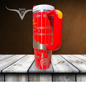 Pu&ograve; includere: Tazza da viaggio in acciaio inox rosso con manico rosso e coperchio bianco. La tazza ha un design con un campo da calcio con un logo di squadra e le parole "City" e "Football".