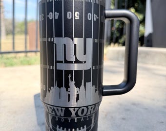 Könnte beinhalten: Schwarzer und silberner Reisebecher mit Griff. Der Becher hat ein Footballfeld-Design mit Yard-Linien und den Buchstaben "NY". Die Skyline von New York und die Worte "NEW YORK FOOTBALL" sind ebenfalls sichtbar.