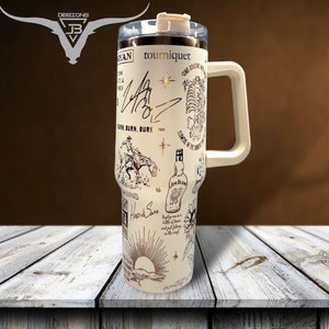 May include: A white stainless steel travel mug with a black and gold design. The mug features a cowboy riding a horse, a bottle of Jack Daniel's whiskey, and the text "tourniquet" and "BURN, BURN, BURN".