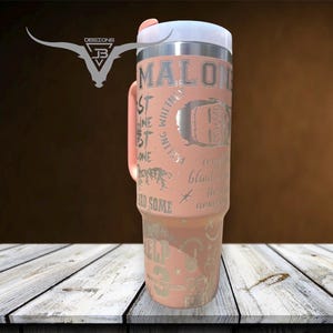 May include: A pink stainless steel travel mug with a white lid and a silver design featuring the text "MALONE" and other words. The mug is on a wooden surface.