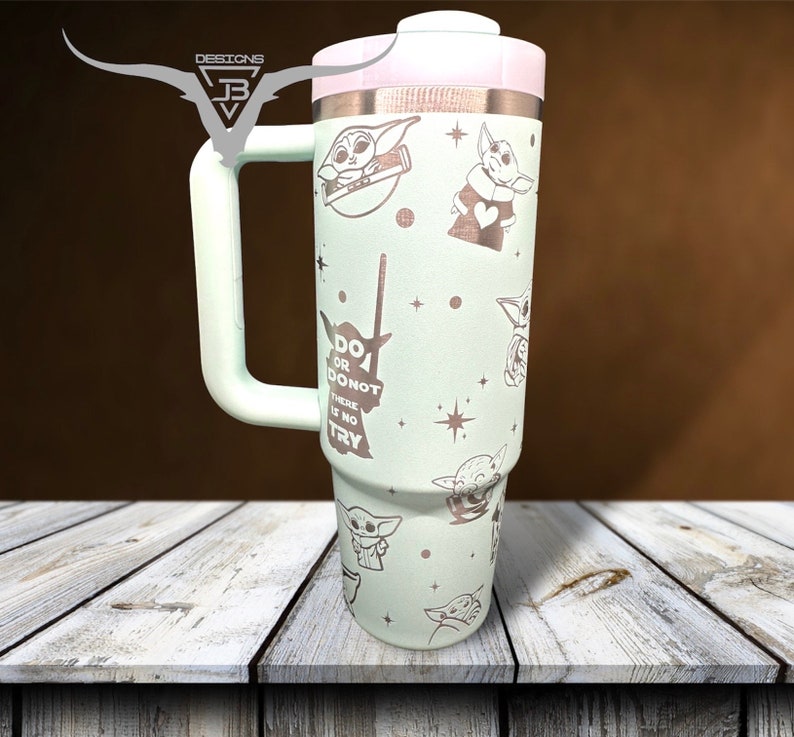 Puede incluir: Una taza de viaje de acero inoxidable verde claro con tapa y asa blancas. La taza presenta un patr&oacute;n repetido de un personaje de dibujos animados con el texto "Do or do not, there is no try".