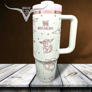Puede incluir: Una taza de viaje de acero inoxidable verde claro con un asa blanca. La taza presenta un patr&oacute;n repetido de un personaje de dibujos animados con el texto "STANLEY" en la taza.