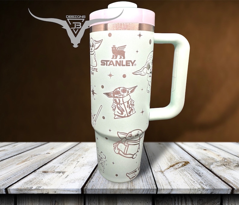 Puede incluir: Una taza de viaje de acero inoxidable verde claro con un asa blanca. La taza presenta un patr&oacute;n repetido de un personaje de dibujos animados con el texto "STANLEY" en la taza.