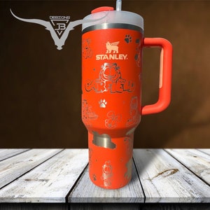 May include: An orange Stanley tumbler featuring Garfield graphics and the Stanley logo. The tumbler has a handle, a white lid, and a straw. The design includes Garfield's name and various cartoon illustrations. The tumbler is on a white wooden surface.