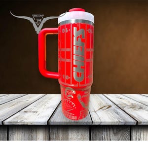 Pu&ograve; includere: Tazza da viaggio in acciaio inox rosso con manico nero e coperchio bianco. La tazza ha un design da campo da calcio con la scritta "CHIEFS" stampata su di essa.