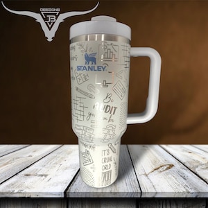 May include: White stainless steel travel mug with a white handle. The mug has a silver design of various office supplies and the text "Be your own audit" and "It's a cruel world". The mug is on a wooden surface.