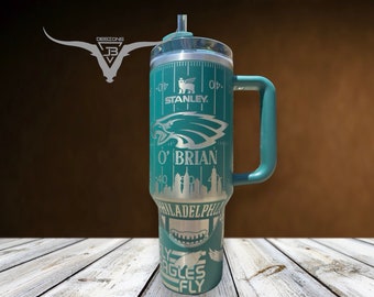May include: A green stainless steel travel mug with a black handle. The mug features a Philadelphia Eagles theme with the team logo, the city skyline, and the words "Fly Eagles Fly". The mug also has a ruler on the side with the words "Stanley" and "O'Brian".