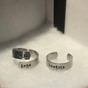 May include: Two silver-colored adjustable rings. One ring has the word "saja" on the top and "boys" on the bottom. The other ring has the word "huntrix" on it. The rings are displayed on a white surface.