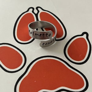 以下が含まれることがあります： 「palmetto」と「foxes」が刻まれた銀色の金属製のリング。リングは赤い足跡のある白い背景の上に置かれています。