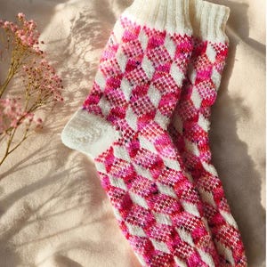 May include: A pair of knitted socks featuring a repeating diamond pattern in shades of bright pink, magenta, and white. The socks have a ribbed cuff at the top and a solid white heel and toe section, crafted with a textured yarn finish.