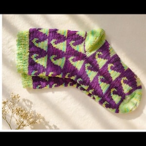 Peut inclure: Paire de chaussettes tricotées avec un motif répété de formes vertes et violettes. Le bord et le bout sont verts. Les chaussettes sont pliées et posées sur une surface blanche.