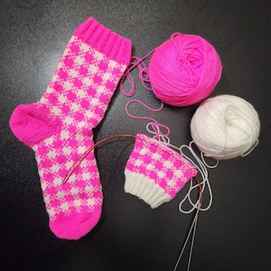 Puede incluir: Un calcetín rosa y blanco con patrón de rombos parcialmente terminado, con agujas de tejer, hilo y una bola de hilo rosa. El calcetín está sobre una superficie oscura, con un segundo calcetín en proceso.