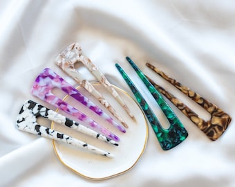 May include: A set of six hair clips in various colors and patterns, including black and white, purple, brown, and green. The clips are arranged on a small white plate with a gold rim.