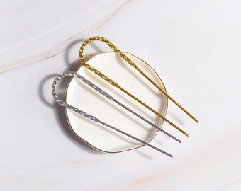 May include: Two hair pins on a white ceramic dish. One hair pin is gold and the other is silver.