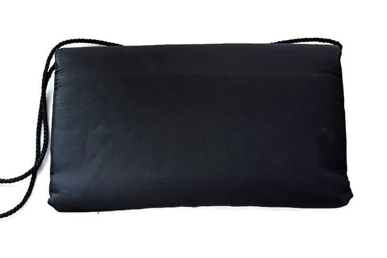 Puede incluir: Un bolso de mano rectangular negro con una correa de cuerda negra. El bolso est&aacute; hecho de un material liso, posiblemente similar al sat&eacute;n. La correa est&aacute; sujeta a las esquinas superiores del bolso.