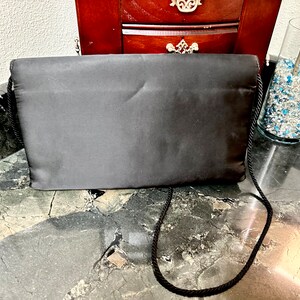 Puede incluir: Bolso de mano de sat&eacute;n negro con una correa para el hombro de cuerda. El bolso rectangular se exhibe sobre una superficie de m&aacute;rmol. El bolso es de color negro liso y tiene un dise&ntilde;o sencillo y elegante. Un jarr&oacute;n de cristal con piedras decorativas azules y transparentes est&aacute; en el fondo.