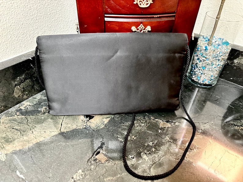 Puede incluir: Bolso de mano de sat&eacute;n negro con una correa para el hombro de cuerda. El bolso rectangular se exhibe sobre una superficie de m&aacute;rmol. El bolso es de color negro liso y tiene un dise&ntilde;o sencillo y elegante. Un jarr&oacute;n de cristal con piedras decorativas azules y transparentes est&aacute; en el fondo.