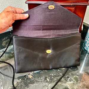 Puede incluir: Bolso de mano tipo sobre de sat&eacute;n negro con una larga correa de cord&oacute;n negro. La solapa se asegura con un bot&oacute;n dorado. El interior es de sat&eacute;n marr&oacute;n oscuro. Un peque&ntilde;o bot&oacute;n decorativo est&aacute; en la solapa.