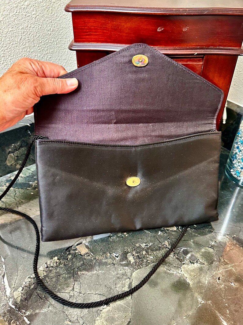 Puede incluir: Bolso de mano tipo sobre de sat&eacute;n negro con una larga correa de cord&oacute;n negro. La solapa se asegura con un bot&oacute;n dorado. El interior es de sat&eacute;n marr&oacute;n oscuro. Un peque&ntilde;o bot&oacute;n decorativo est&aacute; en la solapa.