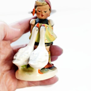Puede incluir: Figura vintage de una ni&ntilde;a con dos gansos. La ni&ntilde;a lleva un pa&ntilde;uelo rojo, un vestido amarillo con lunares rojos y botas marrones. Los gansos son blancos con picos y patas naranjas. La figura est&aacute; sobre una peque&ntilde;a base ovalada.
