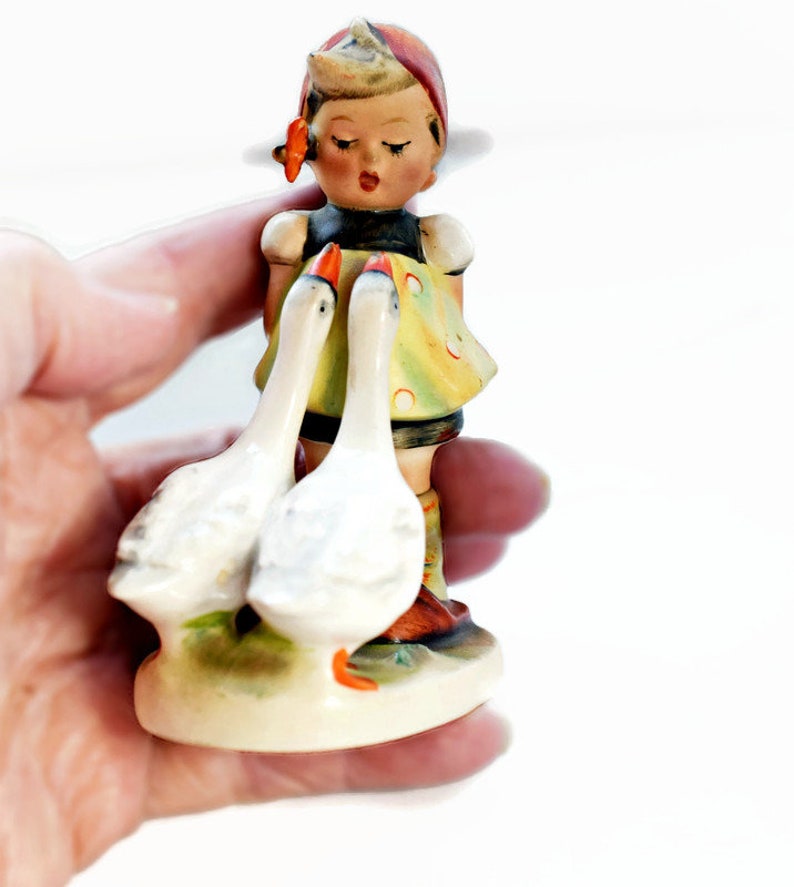 Puede incluir: Figura vintage de una ni&ntilde;a con dos gansos. La ni&ntilde;a lleva un pa&ntilde;uelo rojo, un vestido amarillo con lunares rojos y botas marrones. Los gansos son blancos con picos y patas naranjas. La figura est&aacute; sobre una peque&ntilde;a base ovalada.
