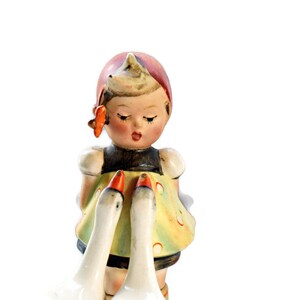 Puede incluir: Figura de porcelana vintage de una ni&ntilde;a con un vestido amarillo con detalles rojos, ribete negro y gorro rojo. Dos gansos blancos con picos naranjas est&aacute;n frente a ella. Est&eacute;tica cl&aacute;sica y nost&aacute;lgica.