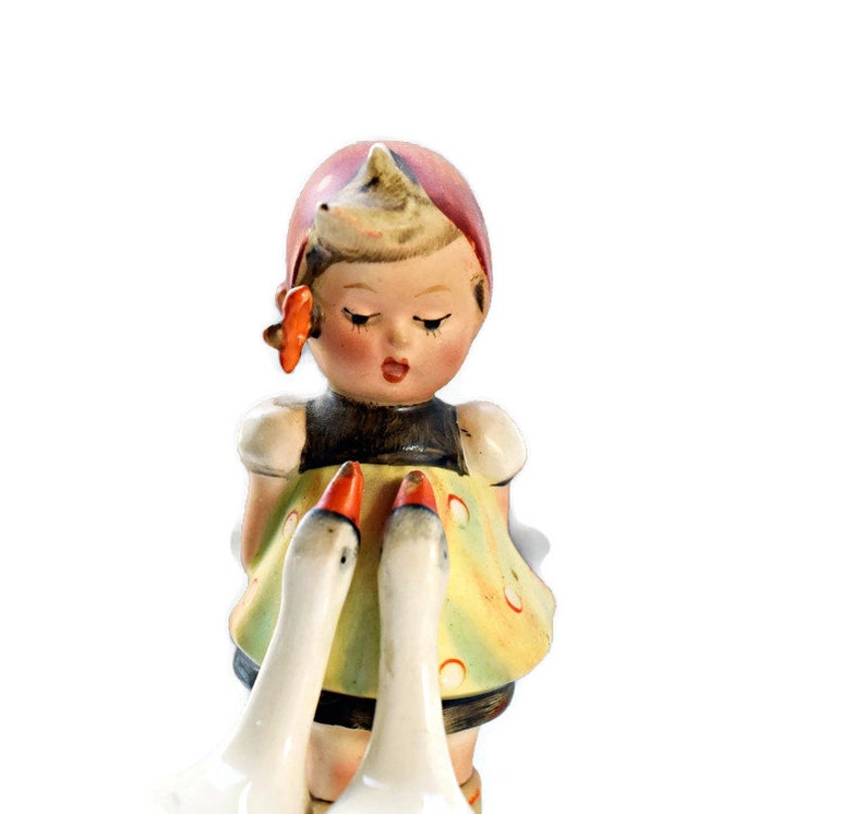 Puede incluir: Figura de porcelana vintage de una ni&ntilde;a con un vestido amarillo con detalles rojos, ribete negro y gorro rojo. Dos gansos blancos con picos naranjas est&aacute;n frente a ella. Est&eacute;tica cl&aacute;sica y nost&aacute;lgica.