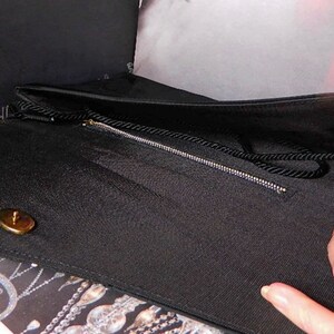 Puede incluir: Bolso de mano negro con cierre y cremallera dorados. El bolso tiene una tela texturizada y un asa negra en forma de cuerda. El interior es negro. La palabra "THE" es visible en una caja negra en el fondo.