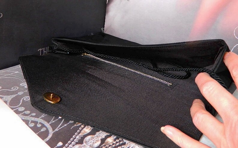 Puede incluir: Bolso de mano negro con cierre y cremallera dorados. El bolso tiene una tela texturizada y un asa negra en forma de cuerda. El interior es negro. La palabra "THE" es visible en una caja negra en el fondo.