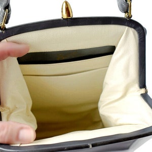 Puede incluir: Bolso vintage negro con cierre y herrajes dorados. El interior es de color crema claro con m&uacute;ltiples compartimentos. El bolso est&aacute; abierto, revelando el interior. El asa es negra y est&aacute; sujeta con herrajes dorados.