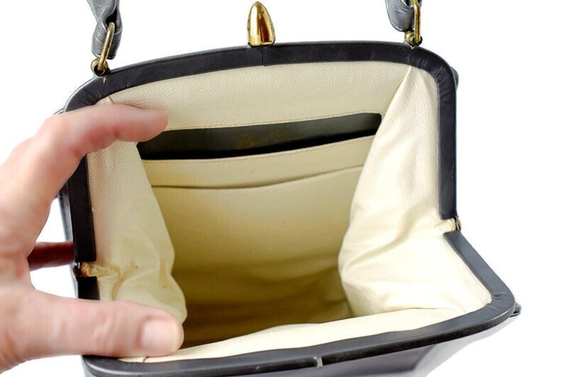 Puede incluir: Bolso vintage negro con cierre y herrajes dorados. El interior es de color crema claro con m&uacute;ltiples compartimentos. El bolso est&aacute; abierto, revelando el interior. El asa es negra y est&aacute; sujeta con herrajes dorados.