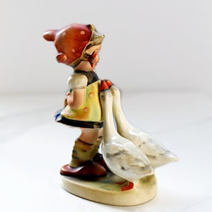 Puede incluir: Figura vintage de un ni&ntilde;o con sombrero rojo de lunares blancos y vestido amarillo, interactuando con dos gansos blancos. La figura tiene un dise&ntilde;o detallado pintado a mano, con una base color crema.