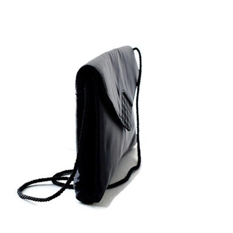 Puede incluir: Bolso de mano negro con cierre de solapa y detalle tejido. El bolso tiene una larga correa de cuerda negra. El bolso est&aacute; hecho de un material liso y brillante.