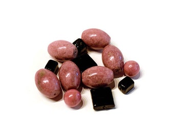 May include: A collection of pink and black gemstone beads. The pink beads are oval and round, while the black beads are square and rectangular. The beads are scattered on a white surface.