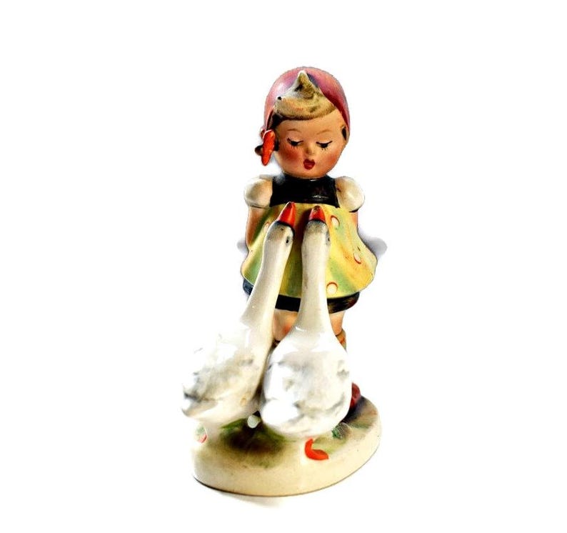 Puede incluir: Una figura vintage de una ni&ntilde;a con un vestido amarillo con detalles rojos, de pie entre dos gansos blancos. La figura tiene un acabado brillante y est&aacute; sobre una peque&ntilde;a base ovalada.