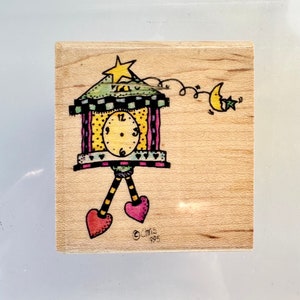 May include: A wooden stamp with a whimsical illustration of a clock tower with a yellow face, a star on top, and a crescent moon with a star on the side. The clock tower has a green roof and pink and yellow stripes. The stamp is signed "Chris 1995".