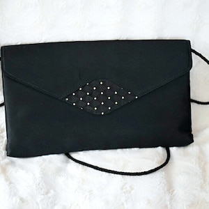 Puede incluir: Un bolso de mano negro con un adorno en forma de diamante con lunares blancos. El bolso tiene un cierre de solapa y una correa de cord&oacute;n negro. Descansa sobre una superficie blanca y esponjosa.