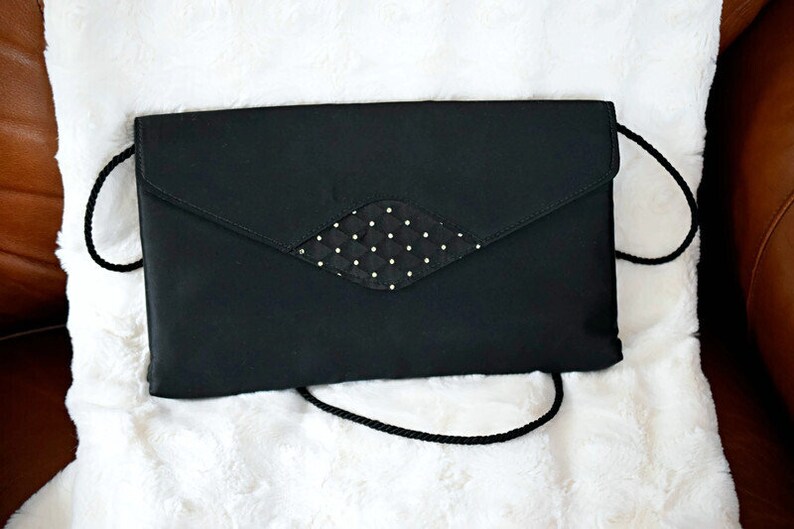 Puede incluir: Un bolso de mano negro con un adorno en forma de diamante con lunares blancos. El bolso tiene un cierre de solapa y una correa de cord&oacute;n negro. Descansa sobre una superficie blanca y esponjosa.