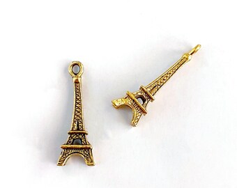 May include: Two gold-colored Eiffel Tower charms. One charm is upright, and the other is angled. The charms have detailed architectural features, including the tower's lattice structure and arched base. The charms are likely for jewelry making or crafting.