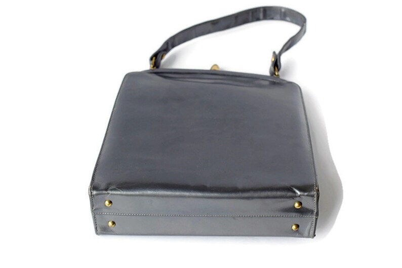 Puede incluir: Un bolso rectangular vintage de color gris oscuro. El bolso tiene un acabado brillante, un asa superior y herrajes dorados. La base del bolso presenta cuatro peque&ntilde;os pies dorados.