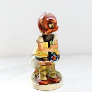 Puede incluir: Figura de cer&aacute;mica vintage de una ni&ntilde;a, vista por detr&aacute;s. Lleva un vestido amarillo con corpi&ntilde;o negro, zapatos marrones y calcetines a rayas. La figura tiene una base en forma de hongo y una cabeza color cobre.
