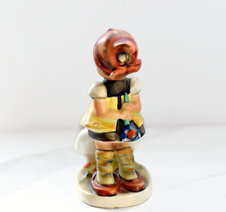 Puede incluir: Figura de cer&aacute;mica vintage de una ni&ntilde;a, vista por detr&aacute;s. Lleva un vestido amarillo con corpi&ntilde;o negro, zapatos marrones y calcetines a rayas. La figura tiene una base en forma de hongo y una cabeza color cobre.