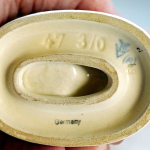 Puede incluir: Base de cer&aacute;mica de figura, color crema, con una abertura ovalada. Estampada con los n&uacute;meros "47 3/0", un emblema azul y la palabra "Germany". La base tiene un borde ligeramente rugoso y texturizado.