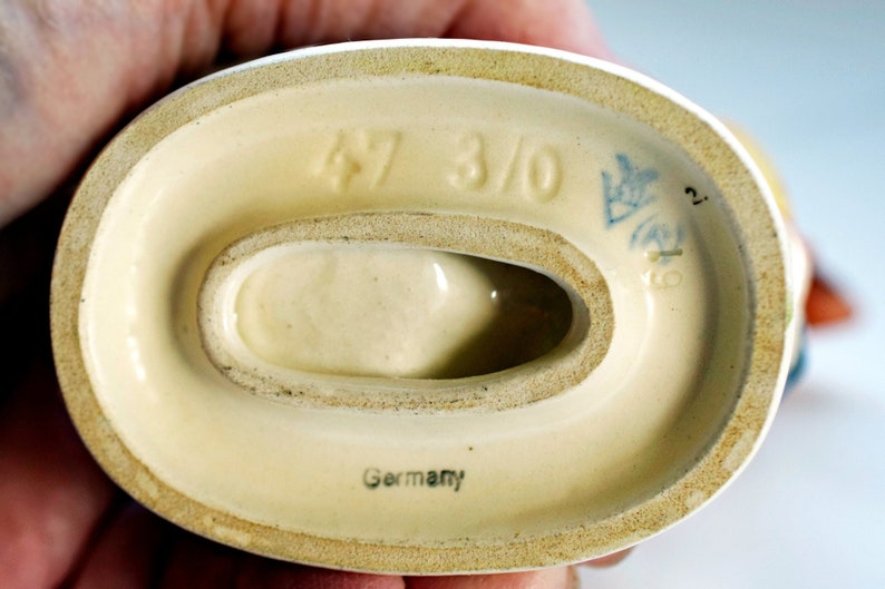 Puede incluir: Base de cer&aacute;mica de figura, color crema, con una abertura ovalada. Estampada con los n&uacute;meros "47 3/0", un emblema azul y la palabra "Germany". La base tiene un borde ligeramente rugoso y texturizado.