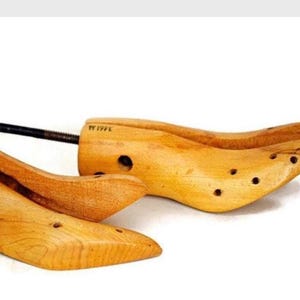 May include: Two wooden shoe stretchers with a natural finish. Each stretcher has a curved shape with a series of small holes and a metal rod with a screw mechanism for adjustment. The shoe stretchers are designed to maintain the shape of shoes.