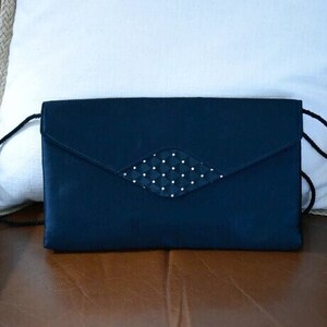 Puede incluir: Un bolso de mano azul marino con un adorno en forma de diamante con lunares blancos. El bolso tiene una correa larga y fina de color negro y un cierre de solapa. El bolso est&aacute; sobre una superficie marr&oacute;n.