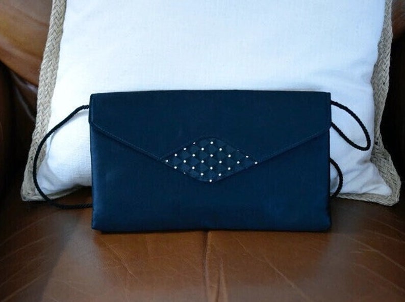 Puede incluir: Un bolso de mano azul marino con un adorno en forma de diamante con lunares blancos. El bolso tiene una correa larga y fina de color negro y un cierre de solapa. El bolso est&aacute; sobre una superficie marr&oacute;n.