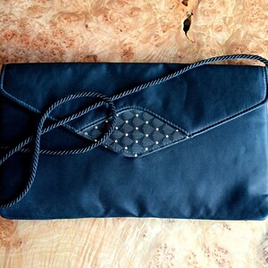 Puede incluir: Un clutch de sat&eacute;n negro con una correa de cuerda a juego. La solapa presenta un panel con dise&ntilde;o de diamantes con peque&ntilde;os adornos dorados. El bolso rectangular es adecuado para ocasiones formales.